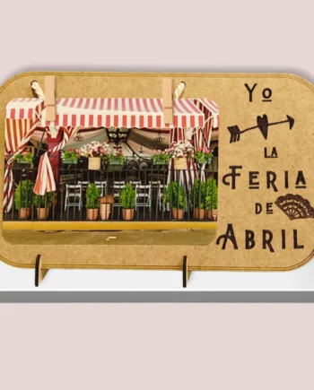 Marcos de fotos de madera con una frase grabada con láser Yo amo la feria de abril. como elementos decorativos tienen un corazón y un abanico. Tiene una foto sujeta con dos pinzas y un cordón de algodón. El portaretratos está puesto con un soporte apoyado en una estanteria